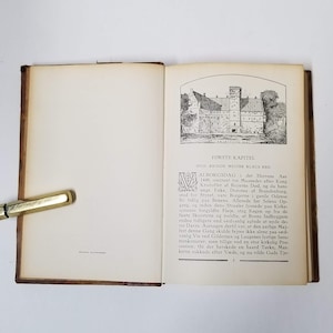 Puede incluir: Un libro antiguo abierto con una cubierta de cuero marr&oacute;n. La p&aacute;gina derecha presenta una ilustraci&oacute;n detallada en blanco y negro de un castillo y texto en idioma extranjero. Una pluma dorada descansa en la p&aacute;gina izquierda.