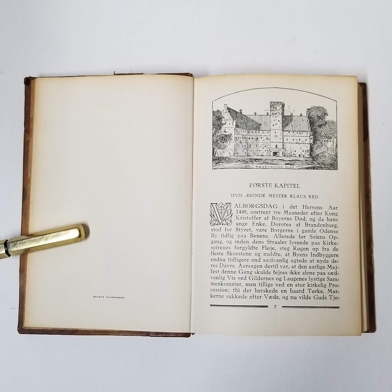 Puede incluir: Un libro antiguo abierto con una cubierta de cuero marr&oacute;n. La p&aacute;gina derecha presenta una ilustraci&oacute;n detallada en blanco y negro de un castillo y texto en idioma extranjero. Una pluma dorada descansa en la p&aacute;gina izquierda.