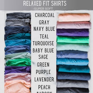 May include: A selection of folded t-shirts in various colours, including charcoal, grey, navy blue, teal, turquoise, baby blue, sage, green, purple, lavender, peach, and maroon. The shirts are labelled with their colour names. RELAXED FIT SHIRTS -SUPER SOFT