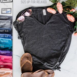 May include: A charcoal gray V-neck t-shirt with short sleeves. The shirt is made of a soft, comfortable fabric and is semi-fitted. The text "RUNS SMALL... SIZE UP FOR RELAXED" is printed on the shirt. The shirt is displayed on a white surface with a pair of brown sandals and pink flowers.