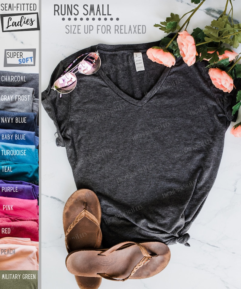 May include: A charcoal gray V-neck t-shirt with short sleeves. The shirt is made of a soft, comfortable fabric and is semi-fitted. The text "RUNS SMALL... SIZE UP FOR RELAXED" is printed on the shirt. The shirt is displayed on a white surface with a pair of brown sandals and pink flowers.