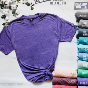 May include: A purple unisex t-shirt with a looser, relaxed fit. The shirt is made of super soft fabric. The image shows a variety of colors available for the shirt, including charcoal, gray frost, navy blue, teal, turquoise, baby blue, sage, peach, maroon, red, royal blue, lavender, and military green.