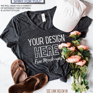 May include: A gray V-neck t-shirt with the text "YOUR DESIGN HERE Free Mock-ups" printed on it. The shirt is laid out on a white marble surface with a white baseball cap, brown sandals, and a bouquet of pink flowers.