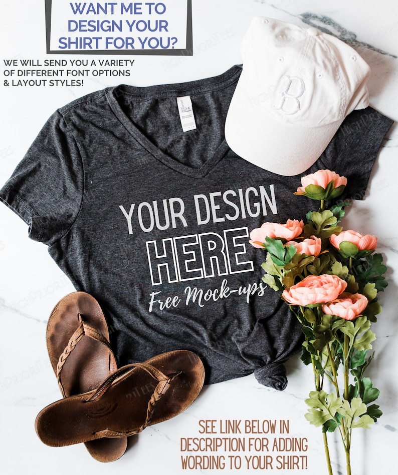 May include: A gray V-neck t-shirt with the text "YOUR DESIGN HERE Free Mock-ups" printed on it. The shirt is laid out on a white marble surface with a white baseball cap, brown sandals, and a bouquet of pink flowers.