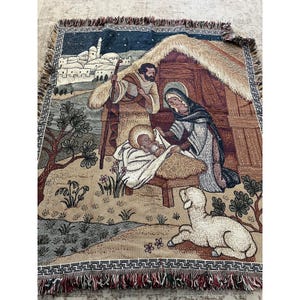 May include: A woven tapestry throw blanket featuring a nativity scene with a central manger, figures of a person standing with a staff and another kneeling, a baby, and a lamb in the foreground, all set against a backdrop of a town and stable.