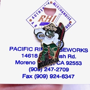 May include: A colorful enamel pin featuring a cartoon frog wearing a crown and holding a hockey stick. The pin is set against a white background with text that reads "PACIFIC RIM GEWORKS" and "BULLFROGS".