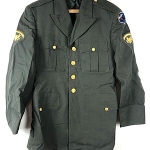 May include: A dark green military uniform jacket with gold buttons and patches on the sleeves. The patches feature a winged eagle and a blue and white insignia.