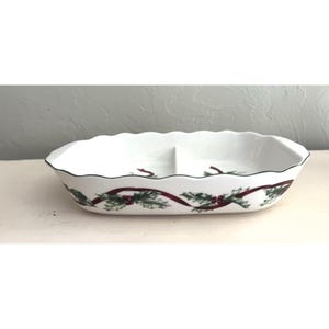 May include: White oval serving dish with two compartments, featuring a scalloped edge and a black trim. The dish is decorated with a festive pattern of red ribbons, green holly leaves, and red berries, perfect for holiday gatherings.
