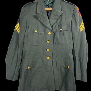 May include: A dark green military uniform jacket with gold buttons and a patch on the right sleeve. The jacket has two pockets on the front and a collar.