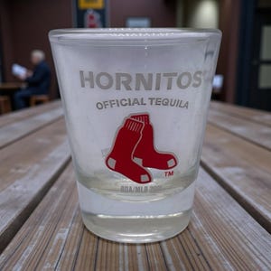 May include: A clear shot glass with the text "Hornitos Official Tequila" and a graphic of two red socks. The shot glass is sitting on a wooden table. The bottom of the glass has the text "BDA/MLB 2021".