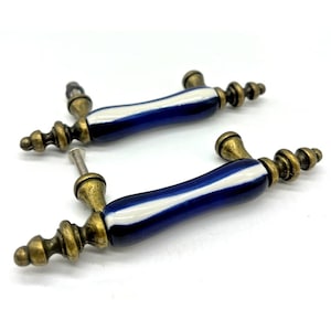May include: Two cabinet handles with a blue and white striped design on the ceramic handle. The ends and hardware are a brass color. The handles are designed for furniture and home decor.