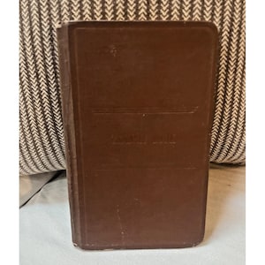 May include: A rectangular brown book with a hard cover. The front cover features a debossed horizontal line design at the top and bottom, with the words "TRAVEL BOOK" embossed in the center. The book has rounded corners and a vertical spine.
