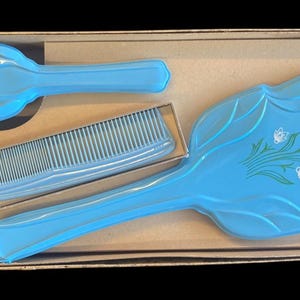 May include: A vintage blue vanity set with a brush, comb, and small hand mirror, all decorated with a floral design. The set is presented in a cardboard box, offering a touch of retro charm.