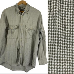 May include: A long-sleeved button-down shirt in a houndstooth pattern, featuring two chest pockets and a collar. The shirt is a muted green and white color, and is hanging on a wooden hanger.
