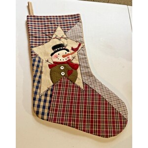 May include: A Christmas stocking featuring a patchwork design of red, blue, and tan plaid fabrics. A large, cream-colored fabric star is appliqued on the front, displaying an embroidered snowman wearing a black hat, red scarf, and a small red bird.