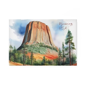 May include: A watercolour painting of Devils Tower in Wyoming, USA. The artwork shows the iconic geological formation in reddish-brown, surrounded by green trees and a blue sky. The text "Wyoming, USA" is written in a script font.