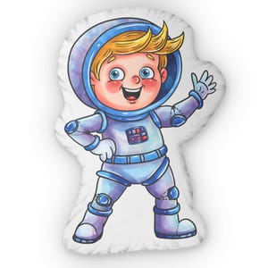 May include: A white pillow featuring a cartoon illustration of a smiling astronaut wearing a blue and white spacesuit. The astronaut has blonde hair and is waving.