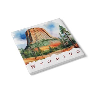 May include: A square, white coaster with a watercolour painting of Devils Tower in Wyoming. The artwork shows the tower in shades of brown and orange, with green trees and a blue sky. The word "WYOMING" is printed in red.