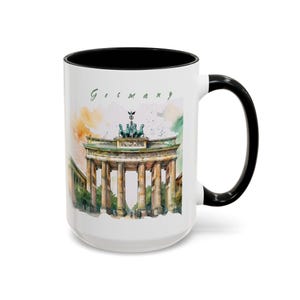 May include: White ceramic mug with a black interior and handle. The mug features a watercolor illustration of the Brandenburg Gate in Berlin, Germany, with the word "Germany" in green script.