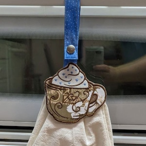 May include: A white kitchen towel with a brown coffee cup design and the text "Coffee is my Love language" embroidered in brown and blue thread. The towel is hanging on a white oven door handle with a blue and white polka dot ribbon.