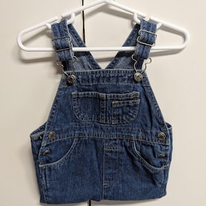 May include: Blue denim overalls for a baby or toddler, hanging on a white hanger. The overalls have pockets on the chest and hips, metal hardware, and button closures. The garment is made of a medium-wash denim fabric.