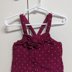 May include: A burgundy corduroy baby dress with white polka dots, hanging on a white hanger. The dress has a ruffled neckline and buttoned straps. The garment is designed for infants and toddlers.