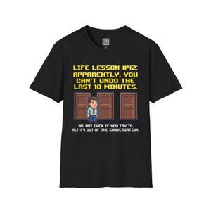 Op de afbeelding: Zwart t-shirt met een pixelachtige afbeelding. De afbeelding toont een persoon voor drie deuren, met de tekst "LIFE LESSON #42: APPARENTLY, YOU CAN'T UNDO THE LAST 10 MINUTES."