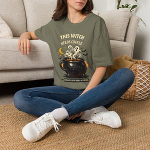 May include: An olive green t-shirt featuring a graphic of a black cauldron with steam rising in the shape of ghosts. The text reads THIS WITCH NEEDS COFFEE and BREWED WITH DARK INTENTIONS, accompanied by a yellow crescent moon and small bats.