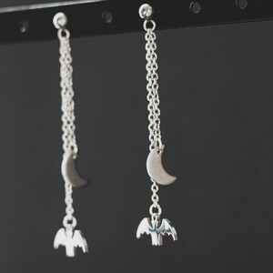 May include: A pair of silver dangle earrings with a crescent moon charm and a bat charm hanging from a silver chain.