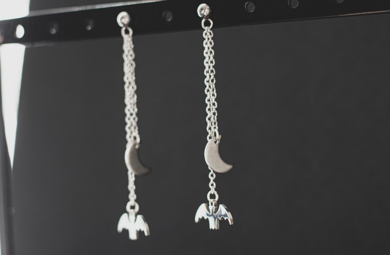 May include: A pair of silver dangle earrings with a crescent moon charm and a bat charm hanging from a silver chain.