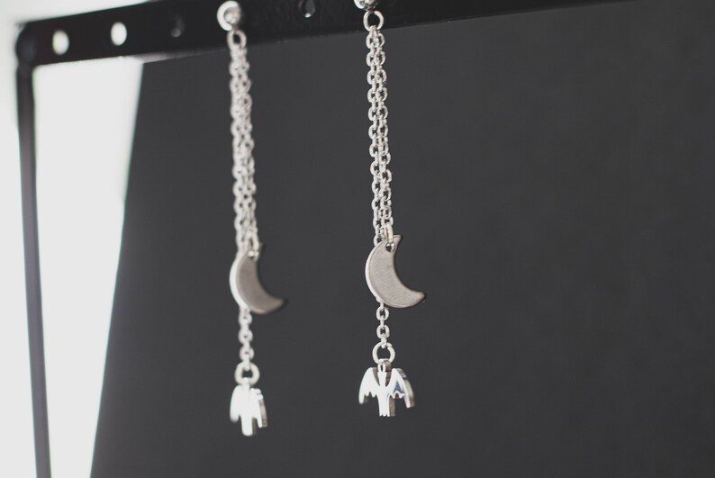 May include: Silver dangle earrings with a crescent moon charm and a bat charm hanging from the bottom.