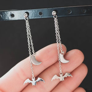 May include: A pair of silver dangle earrings with a crescent moon charm and a bat charm hanging from a chain.