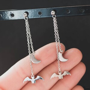 May include: A pair of silver dangle earrings with a crescent moon charm and a bat charm hanging from a chain.