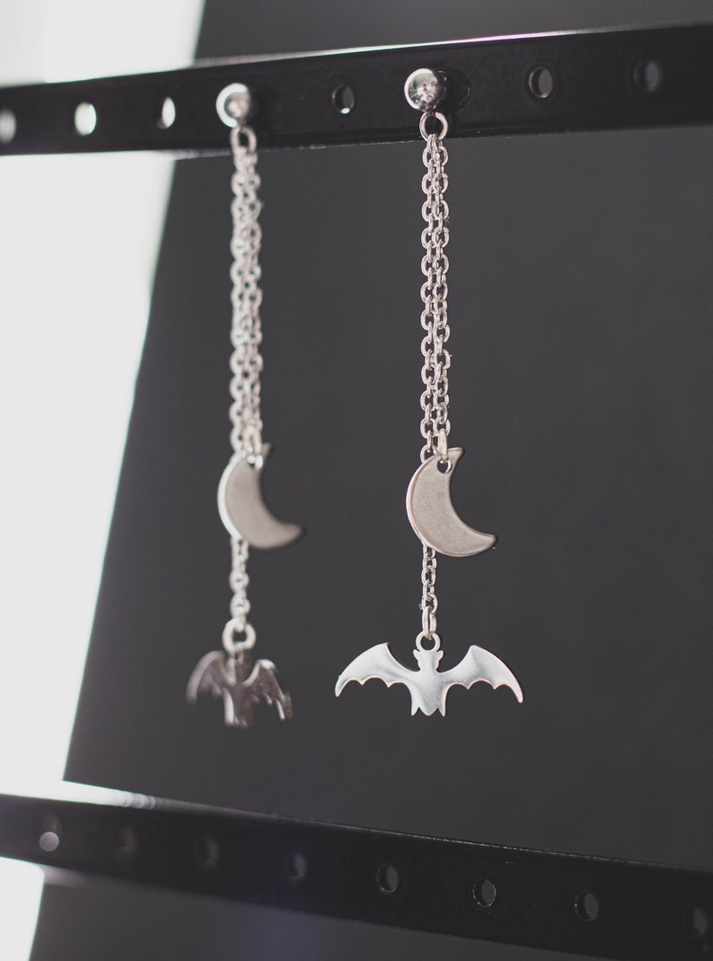 May include: A pair of silver earrings with a crescent moon and bat charm hanging from a silver chain.