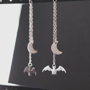 May include: A pair of silver earrings with a crescent moon and bat charm hanging from a silver chain.