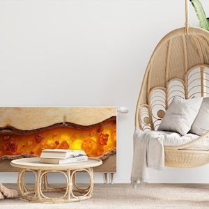 Op de afbeelding: Een witte elektrische radiator met een decoratief paneel van amber agaat. De radiator is gemonteerd op een witte muur in een woonkamer. Een hangende stoel van gevlochten riet met witte kussens staat op de voorgrond.