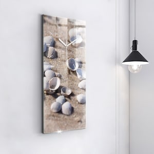 May include: Rectangular wall clock featuring a beach scene with seashells on sand. The clock face has a minimalist design with silver hands and hour markers. The clock is mounted on a white wall, with a black pendant light visible.
