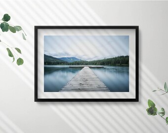 May include: A framed photograph of a wooden jetty extending into a still lake with a mountain range in the background. The water is a light blue and the sky is a pale grey.