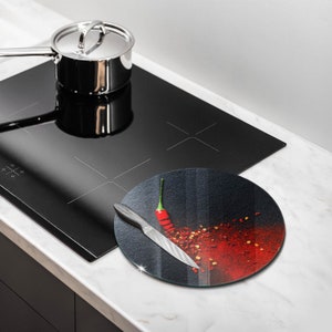 May include: Round glass cutting board with a black background and a red chili pepper and red pepper flakes. A silver knife is on the board.