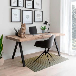 Op de afbeelding: Een licht houten bureau met zwarte metalen poten en een zwarte bureaustoel. Een kat zit op het bureau naast een laptop. Er hangen ingelijste foto's aan de muur achter het bureau.