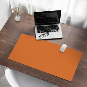 Puede incluir: Una alfombrilla de escritorio naranja con un portátil, gafas y un ratón blanco encima. La alfombrilla de escritorio está sobre un escritorio de madera.