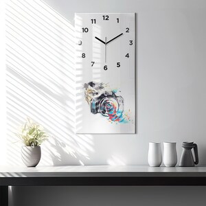 Puede incluir: Un reloj blanco con agujas negras y números negros. La esfera del reloj tiene un diseño de acuarela de una cámara con salpicaduras de pintura de colores. El reloj está colgado en una pared blanca.