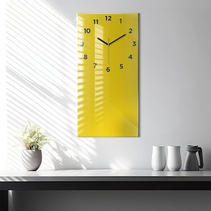 May include: A yellow rectangular clock with black hands and black numbers on a white wall. The clock is hanging above a countertop with a plant and three white mugs.