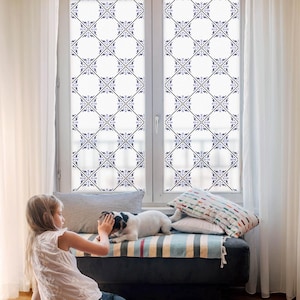 May include: A window with a white frame and a decorative film featuring a repeating pattern of blue and black floral designs. The window is flanked by white curtains. A child and a dog are in front of the window.