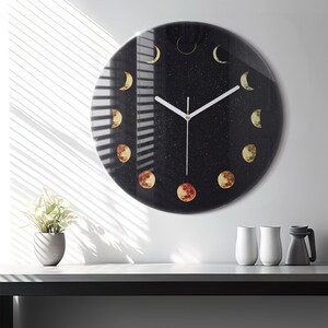 May include: A round clock with a black background and white hands. The clock face features a design of moons in various phases, with a starry sky effect. The clock is made of glass and has a modern design.