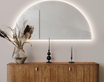 May include: A wooden cabinet with two doors and a half-circle mirror with a light strip around the edge. The mirror is mounted on a white wall above the cabinet. There are two black candle holders with black candles on the cabinet.