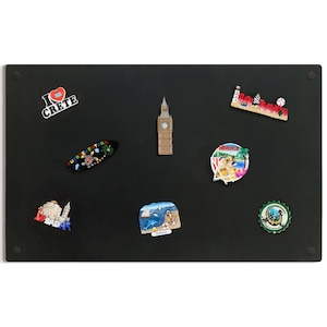 Könnte beinhalten: Eine Sammlung von Souvenir-Magneten auf einer schwarzen Magnettafel. Die Magnete zeigen verschiedene Reiseziele, darunter eine Big Ben-Replik, ein Surfbrett und ein Magnet mit "I Love Crete".