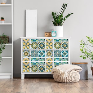 Op de afbeelding: Een witte commode met drie laden versierd met blauwe en gele getegelde stickers met patronen. De commode staat in een kamer met witte planken, planten en een wit frame.
