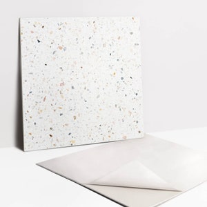 Könnte beinhalten: Eine weiße quadratische Fliese mit einem gesprenkelten Terrazzo-Muster. Die Fliese hat kleine Chips in verschiedenen Farben, darunter Schwarz, Braun, Rosa und Grau.