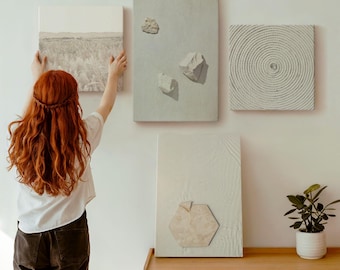 Puede incluir: Una colección de cuatro piezas de arte mural abstracto. Un lienzo presenta un paisaje en escala de grises, mientras que otros muestran diseños texturizados y elementos similares a piedras. El arte se exhibe en una pared blanca, con un mueble de madera y una planta en maceta en primer plano.